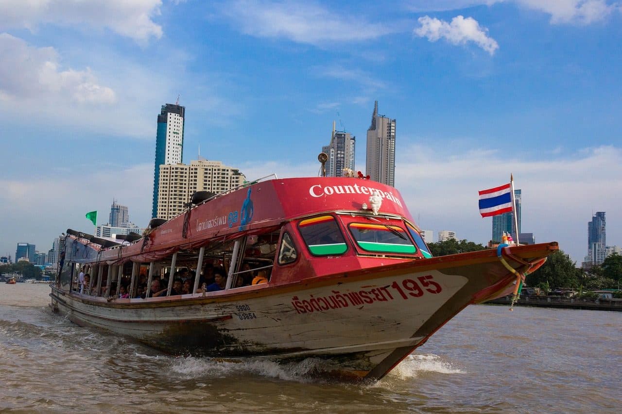 A red river boat travelling along the Chao Phraya with towers on the far bank