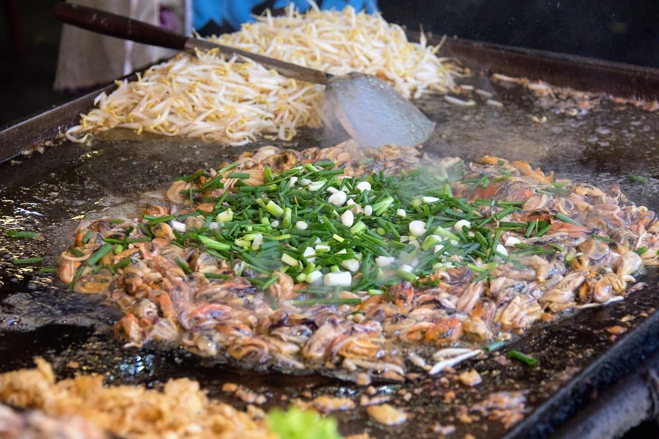 Pad thai being cooked on a large steel griddle with bean sprouts and spring onions
