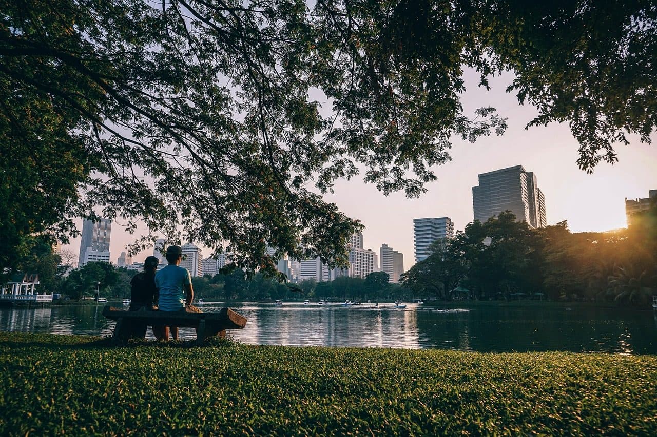 Two people on a bench by a park lake, with Bangkok towers beyond the trees